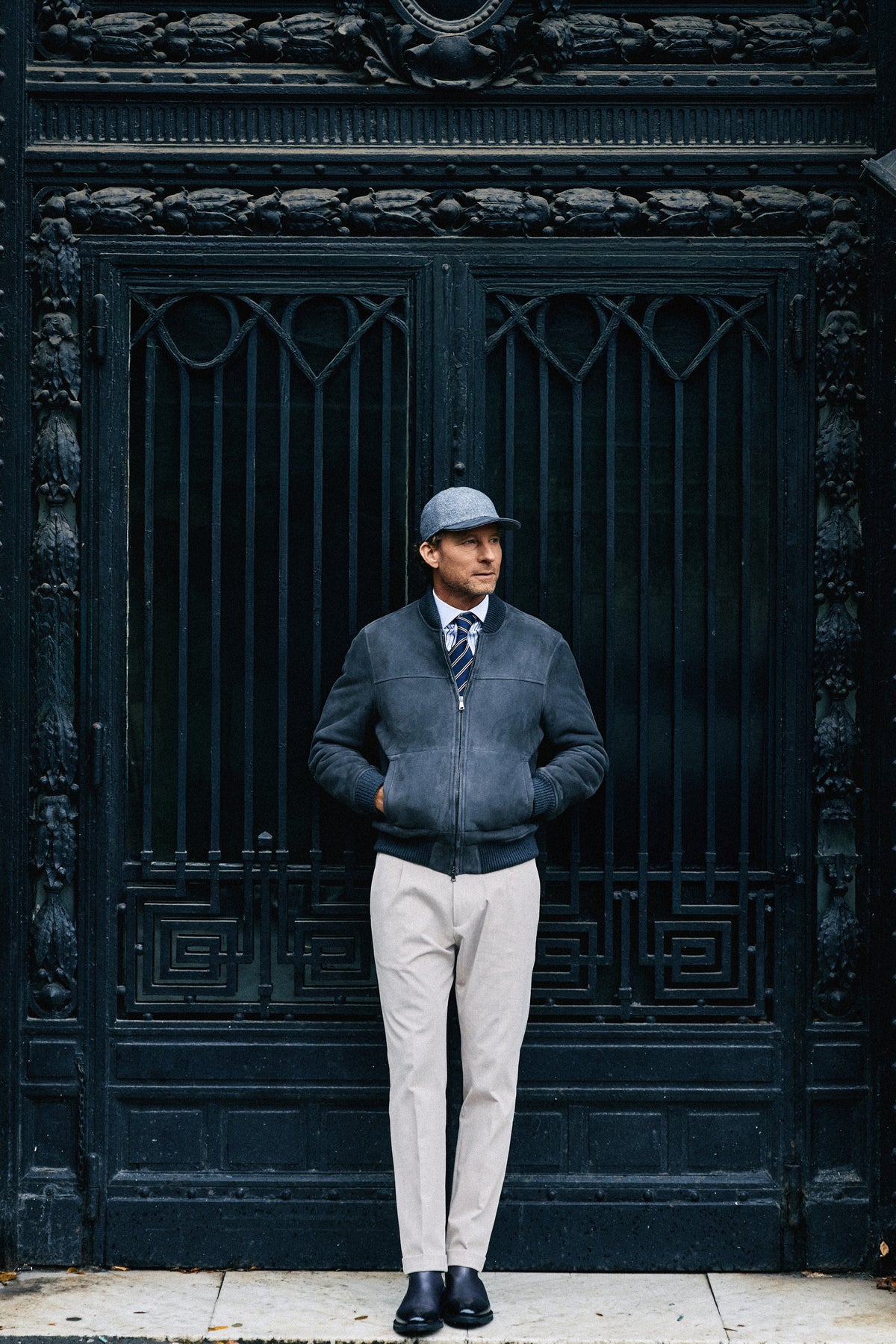 Man in a gray sweater and white pants standing in front of an ornate black door.